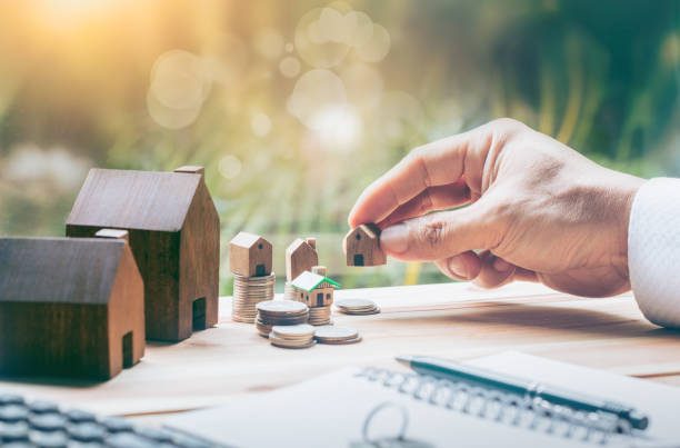 Main plaçant une maison miniature parmi des piles de pièces de monnaie sur un bureau