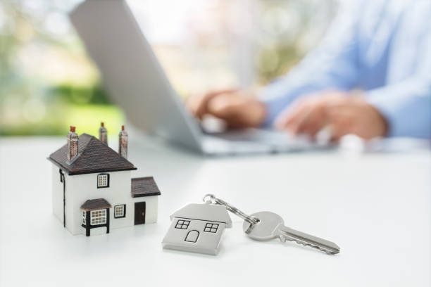 Maison miniature et porte-clés posés sur un bureau, personne travaillant sur un ordinateur portable en arrière-plan